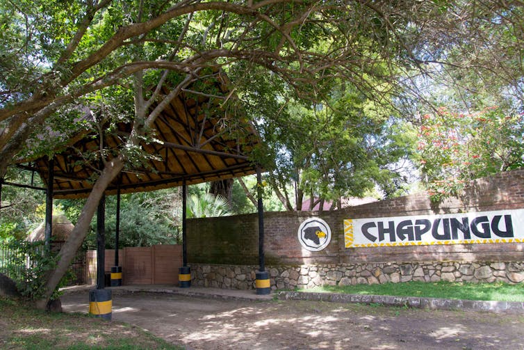 A thatched entrance amid trees, with a large sign on a stone wall reading