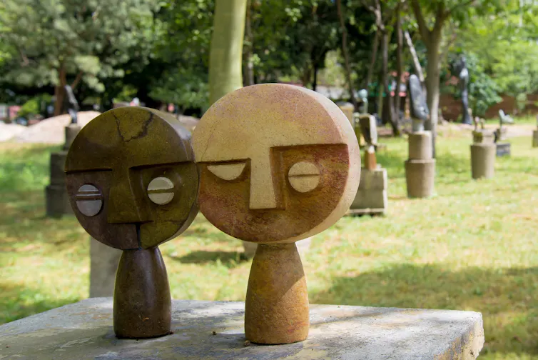 Two round-headed human figures on a plinth, many sculptures in the background.