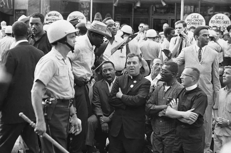 A black and white photo shows four men, two of whom are in clerical suits, kneeling amid a crowd on a sidewalk as a policeman with a baton stands nearby.