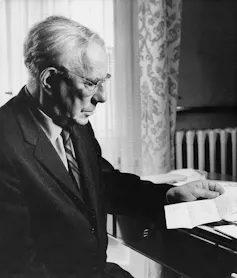A black and white photo of a white-haired man in a suit and tie reading a letter at a desk.