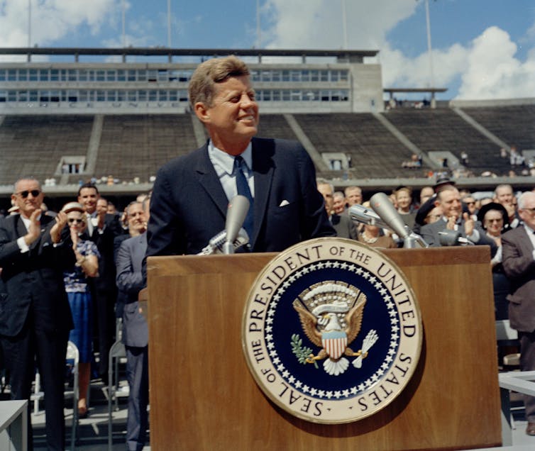 JFK stands at a lectern outdoor in a sports stadium