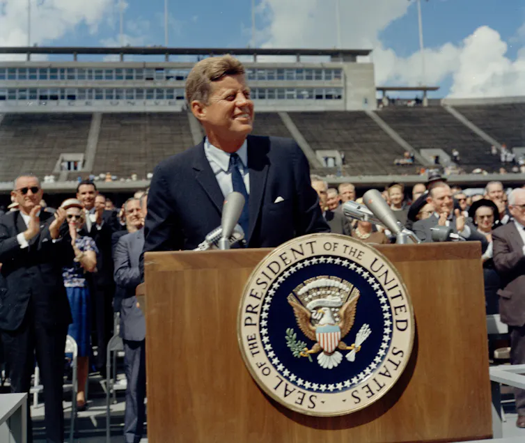 JFK stands at a lectern outdoor in a sports stadium