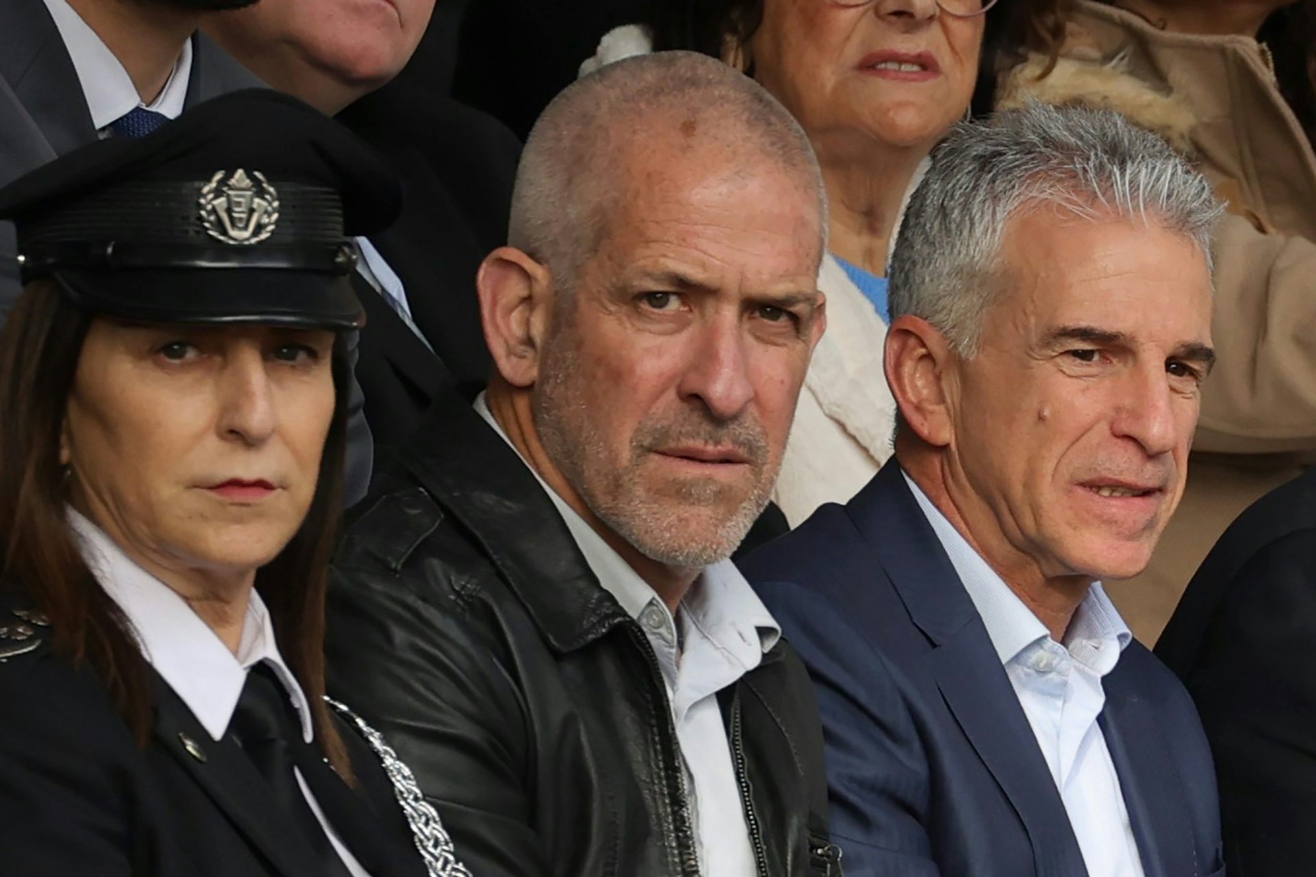 Ronen Bar wearing a leather jacket sat with a woman in security uniform and a man in a suit.