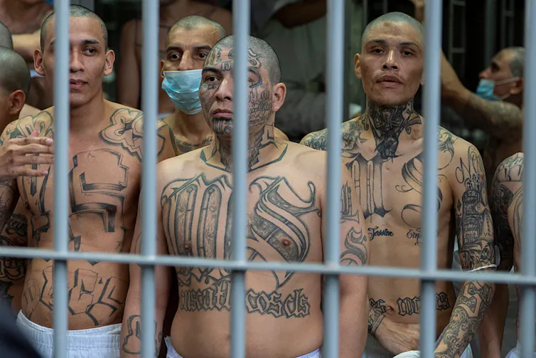 Group of tattooed men stand behind prison bars.