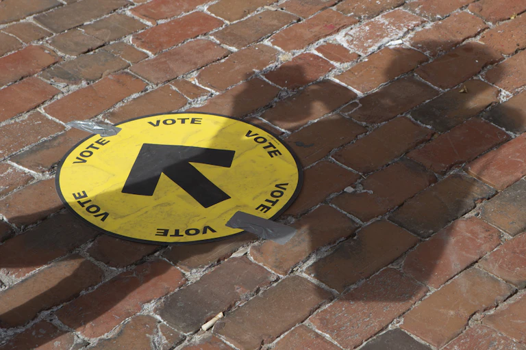 A person'as shadow falls across a yellow circle with arrow on a cobblestoned area, directing people where to vote