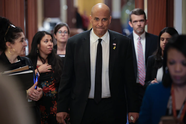 A man with brown skin, a bald head and a black suit with white shirt and black tie smiles and walks in the center of a group of several people who are all dressed formally.