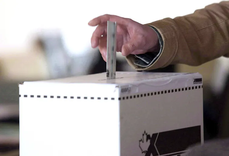 A hand dropping a ballot into a white box with the Elections Canada logo on it