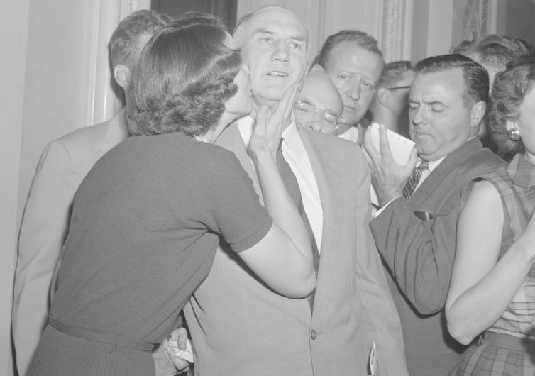 A bald man is kissed on the cheek by a woman, surrounded by other people, in a black and white photo.