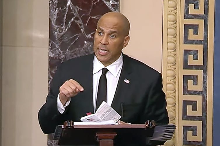 A brown skinned man with a black suit, white shirt and black tie stands at a podium and points his finger toward papers on the podium.