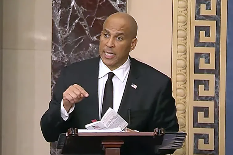 A brown skinned man with a black suit, white shirt and black tie stands at a podium and points his finger toward papers on the podium.