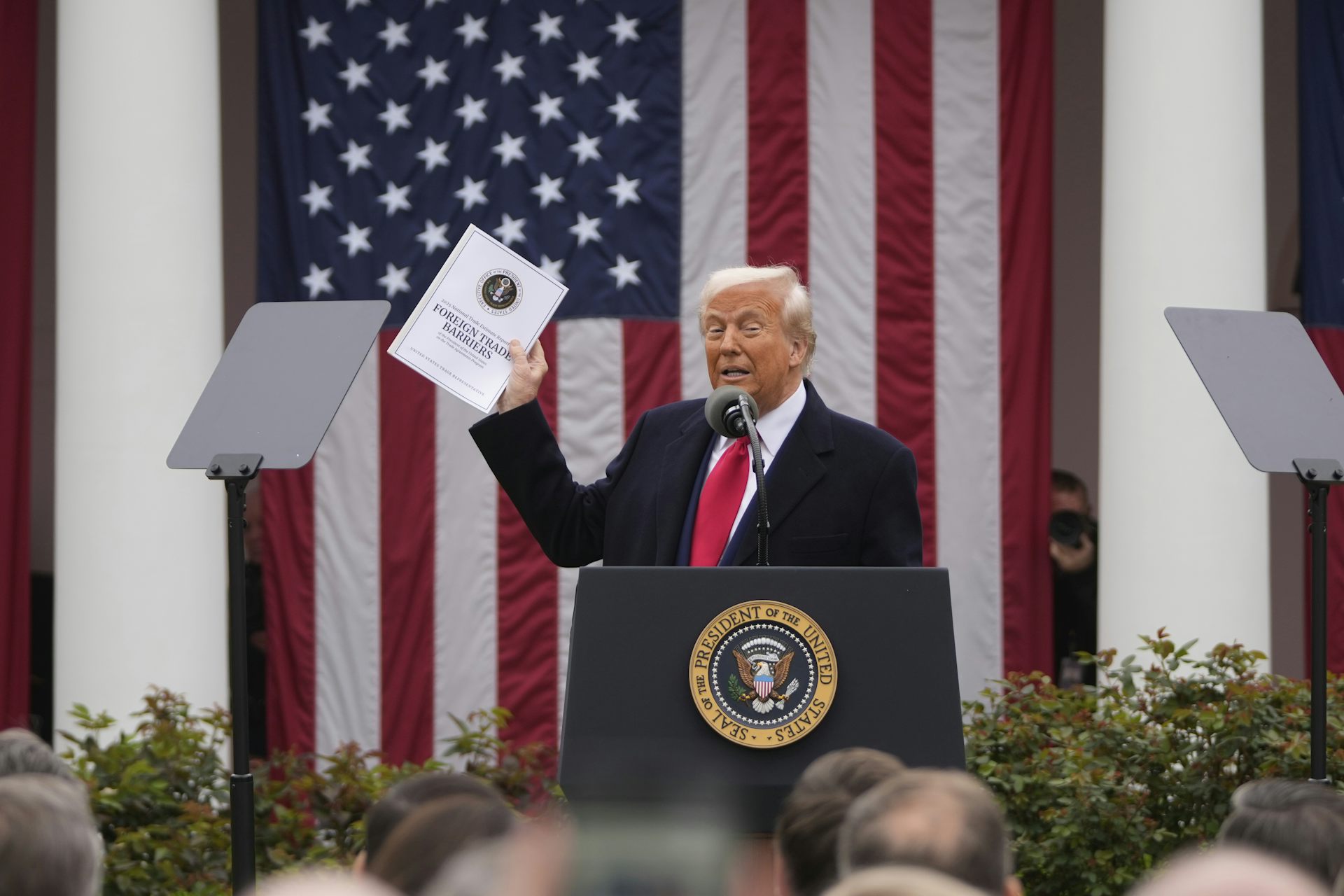 An old man with orange-tinged skin and orange-ish white hair holds up a document that says 'Foreign Trade Barriers' as he stands in front of a giant American flag and behind a presidential lectern.