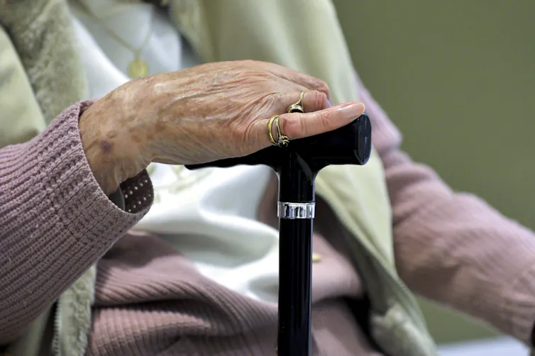 An elderly lady in a pink jumper, with a green towel over her shoulders, and her hand on her walking stick
