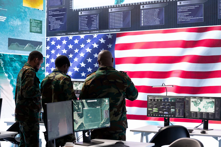Stock phonto of men in military uniform in a secure room with computers and a large US flag.