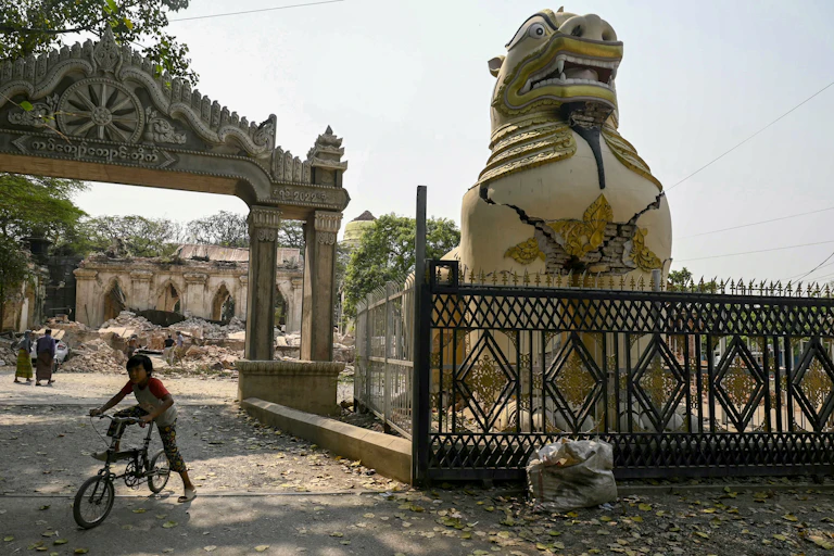Un enfant joue sur un vélo à côté d'une statue de lion bouddhiste fissurée dans la banlieue de Mandalay, le 2 avril 2025, cinq jours après qu'un important tremblement de terre a frappé le centre du Myanmar.