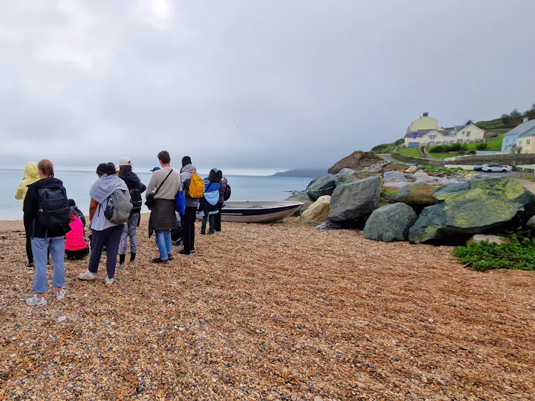 A group of students stood looking out to sea on beach, housing and rocks in background.
