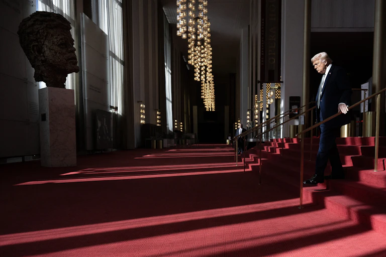 Man in suit descends red carpeted steps with a large bust of a young man facing him.