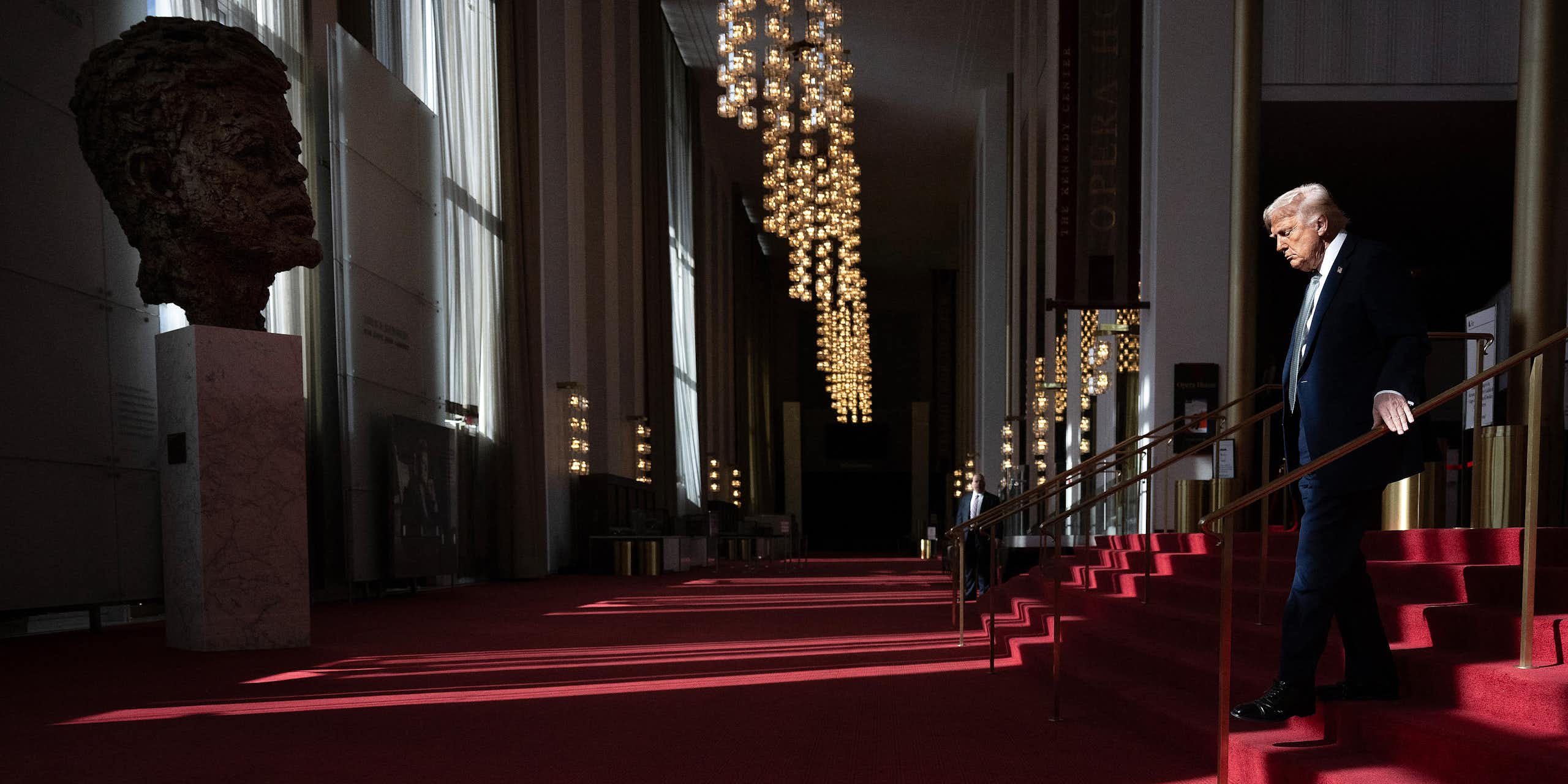 Man in suit descends red carpeted steps with a large bust of a young man facing him.