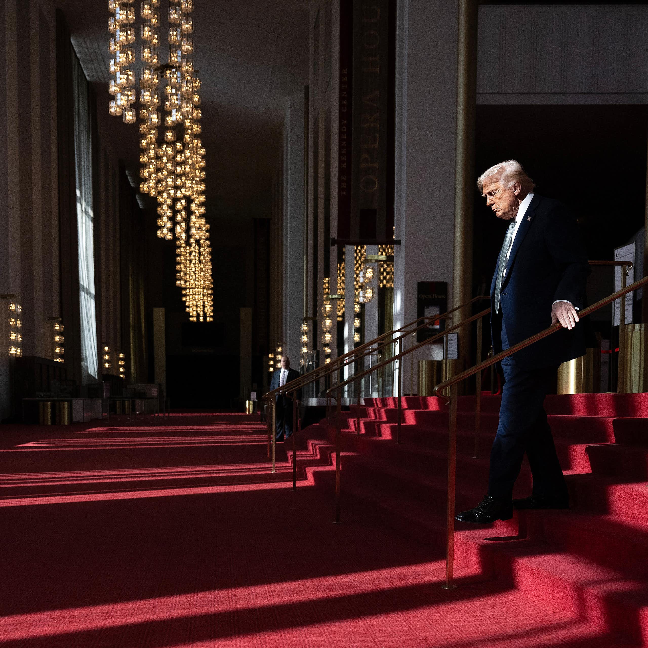 Man in suit descends red carpeted steps with a large bust of a young man facing him.