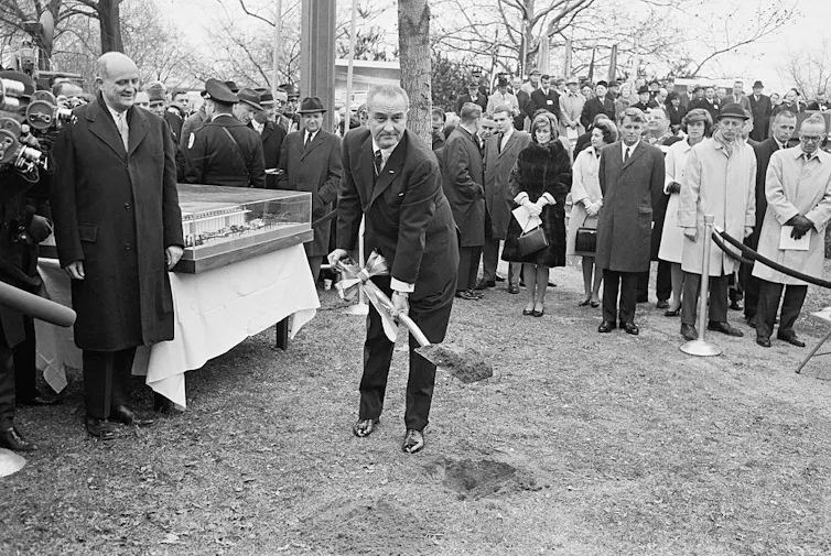 Crowd of people and TV cameras surround man shoveling dirt.