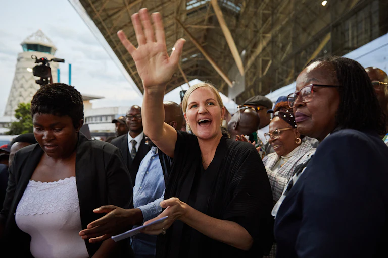 A woman smiles and waves on a stadium grandstand, flanked by African women.
