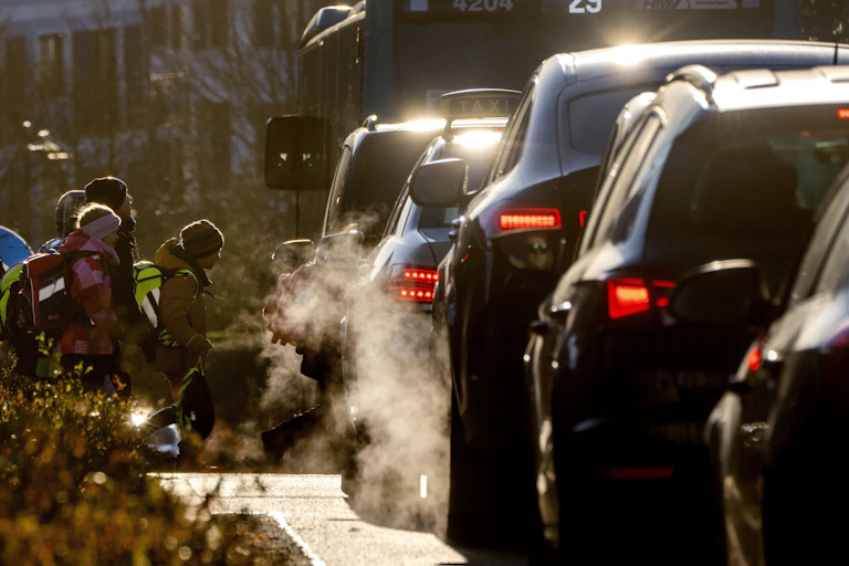 Children walk to school as cars spewing exhaust are on the road next to them.
