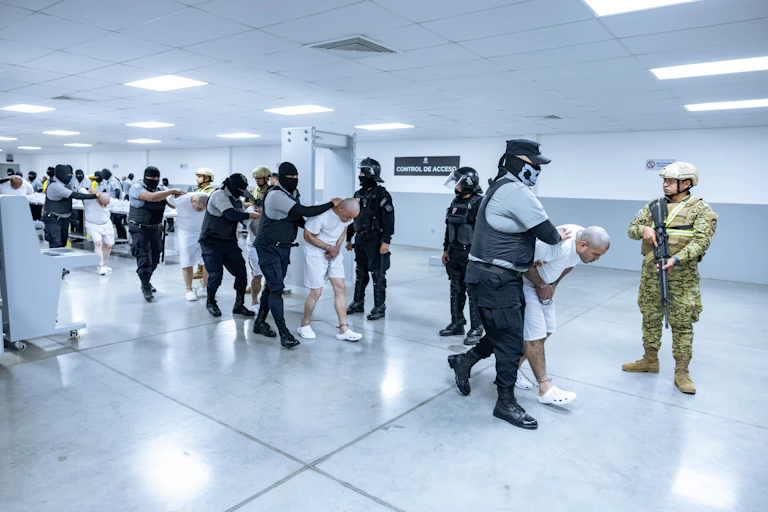 Men wearing white with their heads bent down low are held by men in dark prison uniforms and are led through a white hall.