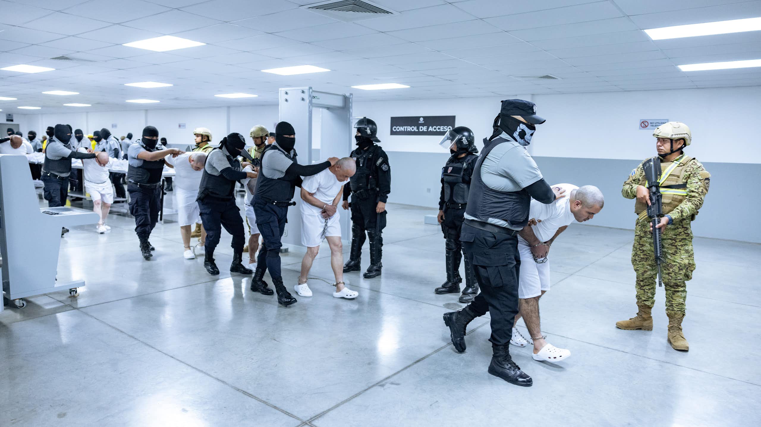 Men wearing white with their heads bent down low are held by men in dark prison uniforms and are led through a white hall.