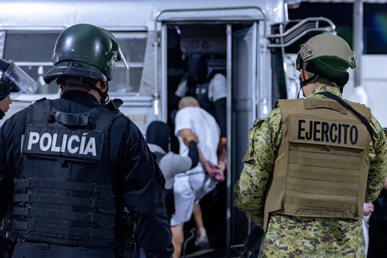 Two people wearing police and army uniforms face men in white who are boarding a bus.