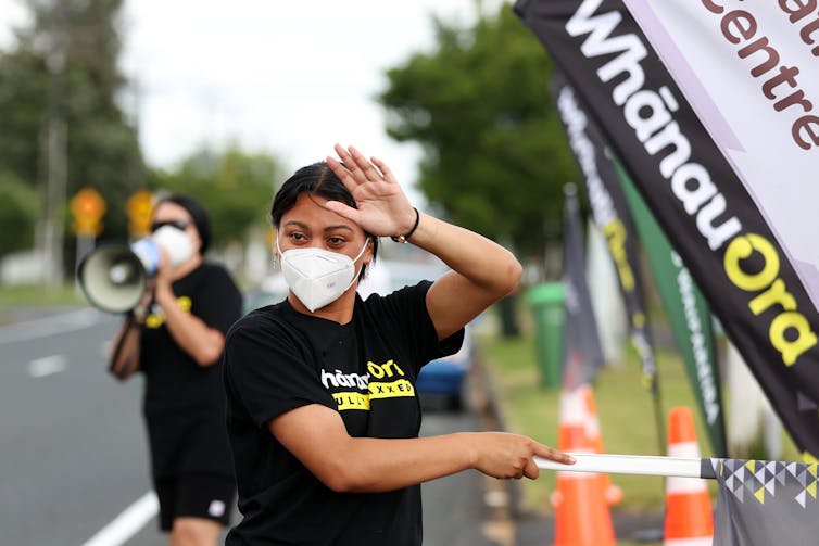 Young Māori woman in face mask waving to traffic next to a Whānau Ora sign during a Covid vaccination drive.