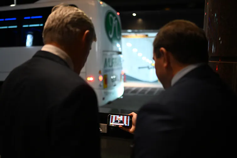 Two men study a smartphone that plays a Donald Trump press conference