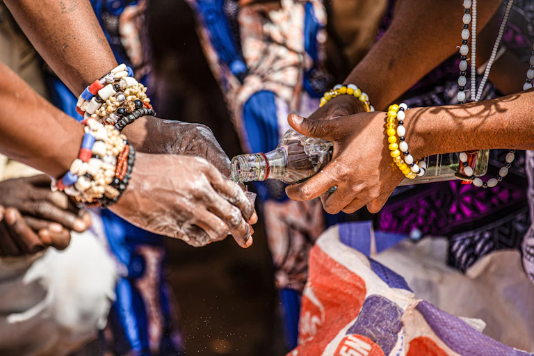 One person's hands holding a bottle, pouring something into the hands of another person. Both people are wearing bead bracelets.