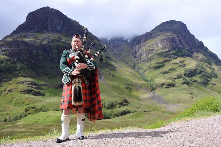 A bagpiper in full ceremonial dress playing bagpipes against striking highlands scenery