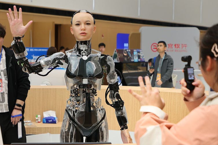 Robot with exposed gears and wiring but with a synthetic, humanlike face and hands raises its right arm as people take photographs.
