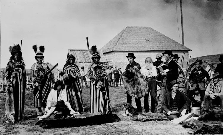 Black and white photo of a group of Indigenous people trading furs with white European settlers