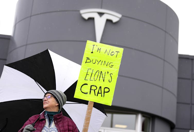 La persona tiene letreros de Neon Yellow Lectura No compro la mierda de Elon frente al juego de Tesla con el logotipo de Tesla que se puede ver sobre el letrero.