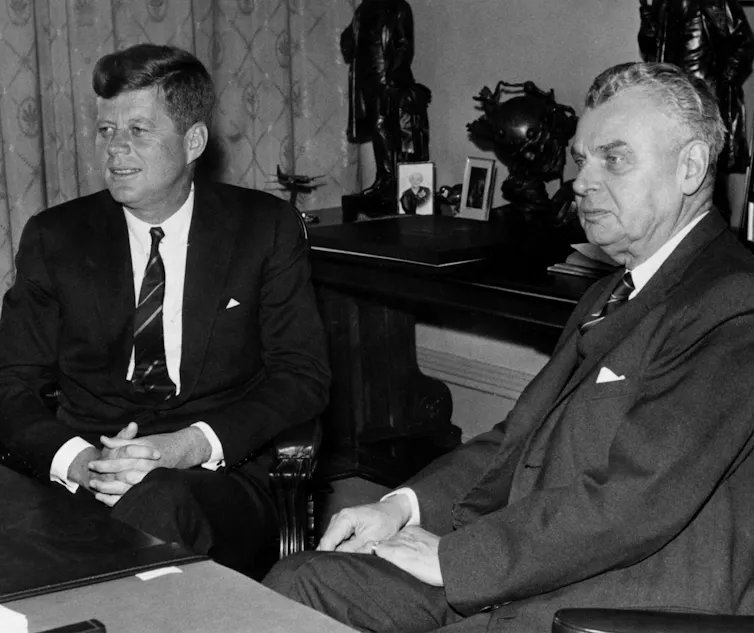 A black-and-white photo shows two men in suits sitting side by side in an office, one a younger dark-haired man and another older, grey-haired man.