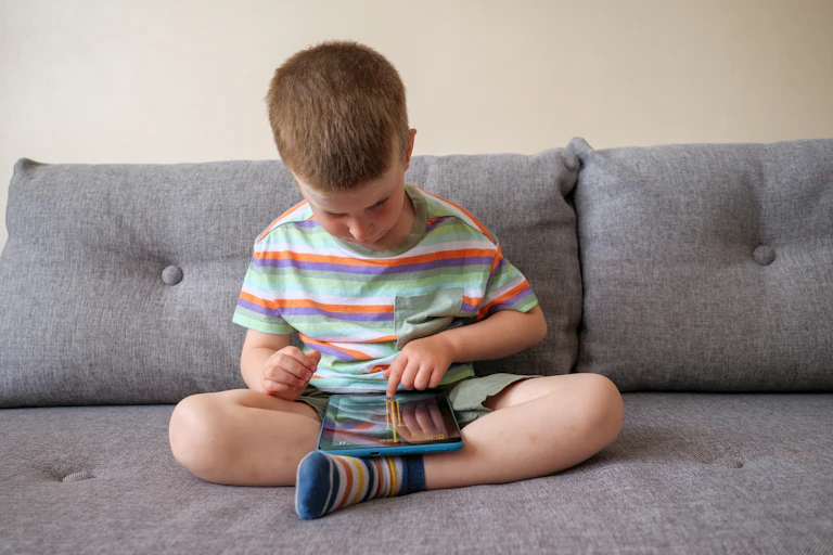 A young boy sits on a couch with a tablet.