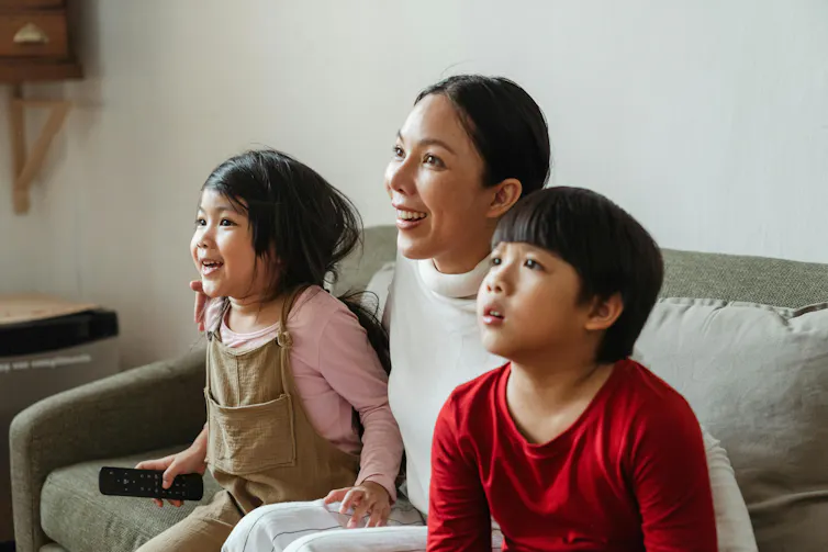 A woman and two young children watch TV on the couch.