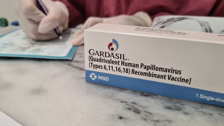 Gloved hands write on a clipboard next to box of the vaccine Gardasil.