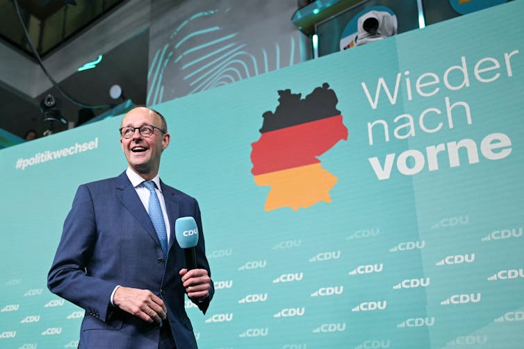 Friedrich Merz holding a microphone in front of a political banner.