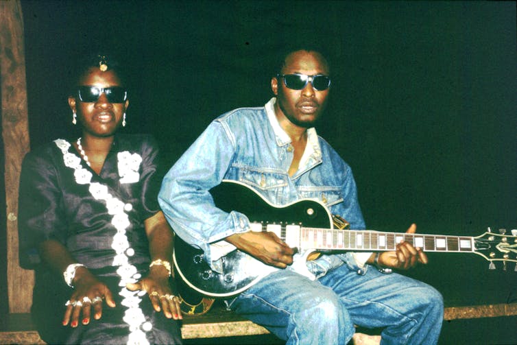 Una joven pareja africana con gafas de sol, la ignorancia de camisas y pantalones y con un vestido blanco y negro, pose de la foto. Sostiene la guitarra.