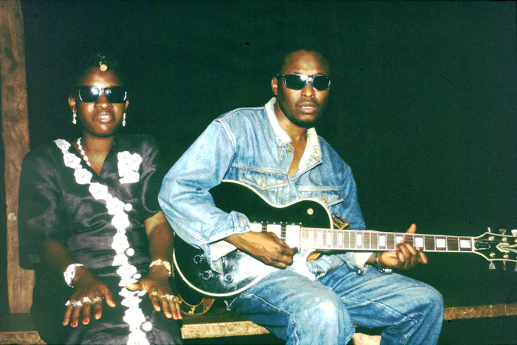 A young African couple in sunglasses, he in denim shirt and pants and she in a black and white dress, pose for a photo. He holds a guitar.