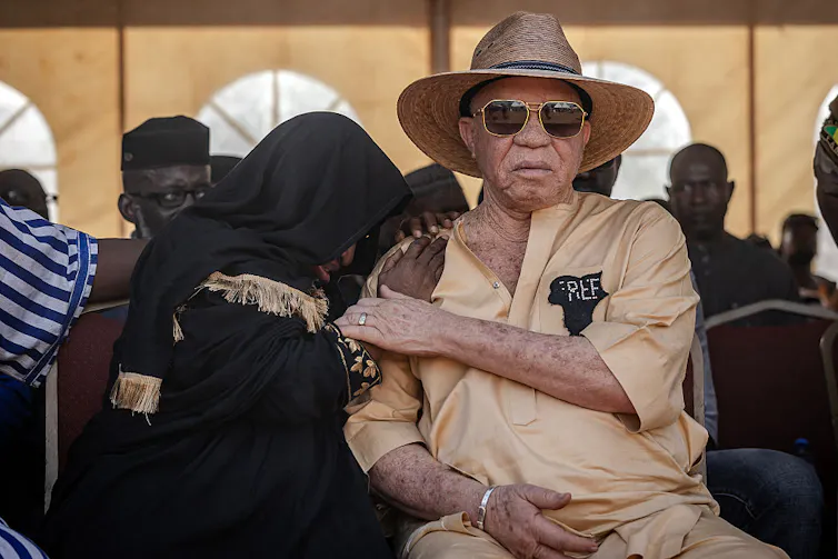 A woman in black mourning dress hides her face on the shoulder of an albino man in sunglasses and a hat, who comforts her.