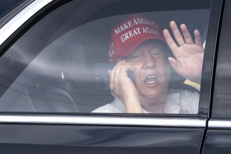 An elderly man with a mobile phone to his ear and wearing red Make America Great Again ballcap waves from the back of a limousine.
