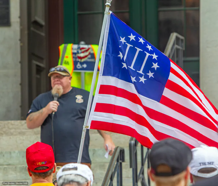 A man addressing a rally, with a flag featuring red stripes and stars arranged in a circle on a blue background, in front of him.