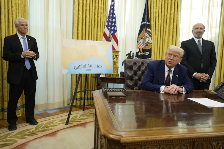 A man with orange-tinged white hair and skin sits at an ornate desk with two men on either side of him. Behind him is a may of the southern United States that reads Gulf of America where the Gulf of Mexico is located.