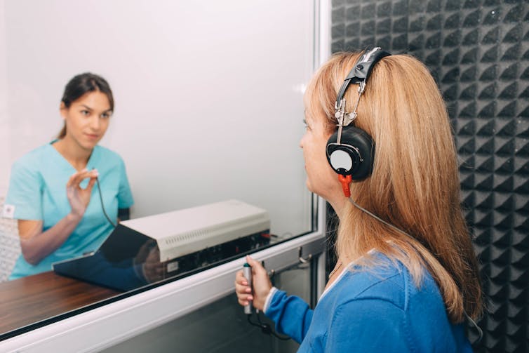 A woman wearing headphones in a booth and an audiologist giving a hearing test