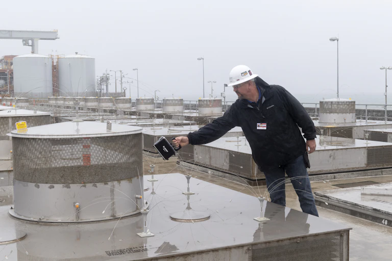 A man in a hard hat holds a piece of electronic equipment near a metal and concrete structure.