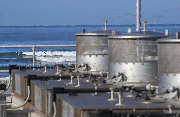 In the foreground, concrete and metal structures are arranged in a line; in the distance, a surfer rides a breaking wave.