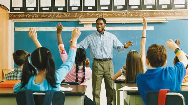 a black male adult stands at the front of a classroom as school-age children sitting at desks raise their hands